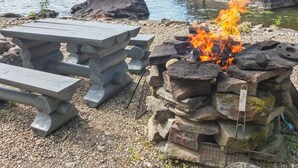 A stone fire pit with a fire burning, surrounded by a wooden picnic table and benches. - Lomasuvas by Interhome (Tuusniemi)