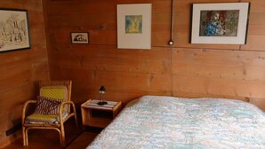 Ein Zimmer mit Holzboden, einem Bett, einem Sessel, einem Nachttisch und gerahmten Bildern an der Wand.