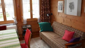 Ein gemütliches Zimmer mit einem gestreiften Sofa, einem kleinen Tisch und einem Stuhl. Es gibt zwei Fenster mit Vorhängen und eine Wandlampe.
