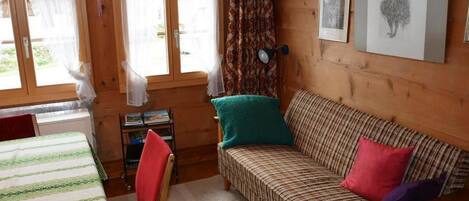 A cozy room with a striped sofa, a small table, and a chair. There are two windows with curtains, and a lamp on the wall.