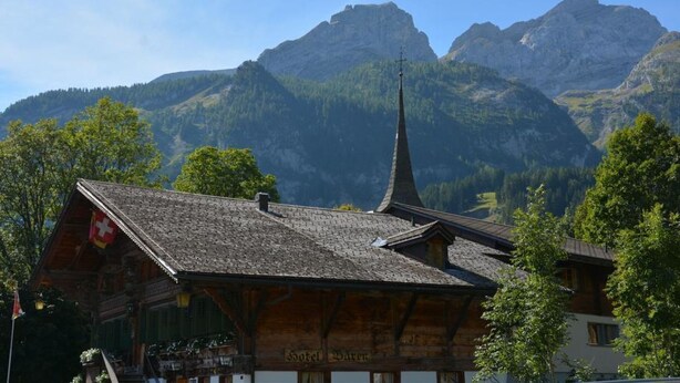Ein traditionelles Holzhaus mit steilem Dach, einem sichtbaren Kirchturm und Bergelandschaft im Hintergrund.