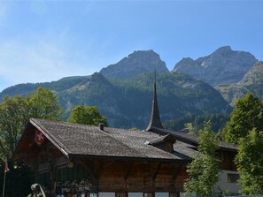 Ein traditionelles Holzhaus mit steilem Dach, einem sichtbaren Kirchturm und Bergelandschaft im Hintergrund.