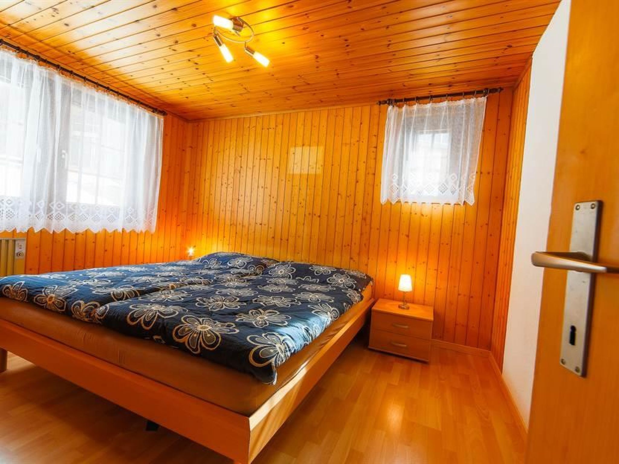 Une chambre avec des murs en bois, un lit recouvert d’une couvre-lit à motifs floraux, deux tables de chevet et un ventilateur de plafond.