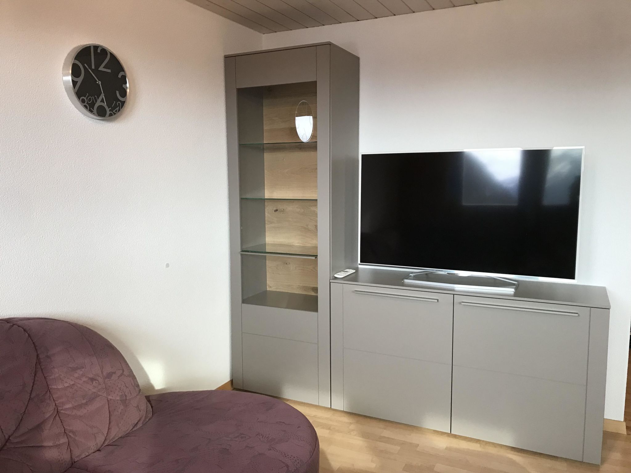 A living room with a grey entertainment unit, a flat-screen TV, and a round wall clock.