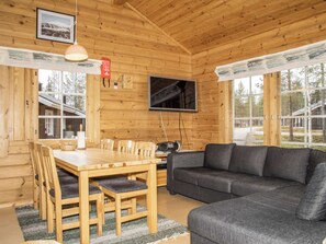 A wooden cabin interior with a dining table, chairs, a sofa, a TV, and a window.