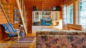 L’intérieur d’un chalet confortable, avec des murs en rondins de bois, un escalier en bois, un canapé, un téléviseur et une bibliothèque.