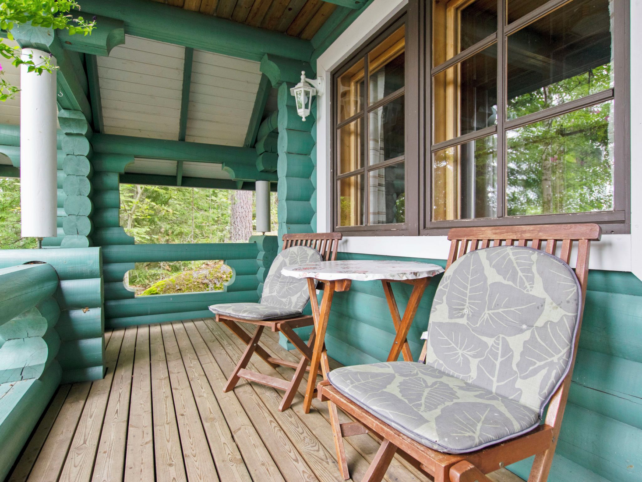 A wooden deck with two wooden chairs and a small table, a window with curtains, and a view of greenery outside.