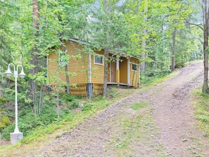 Une petite cabane en bois, accessible par un sentier de terre, et entourée d’arbres.