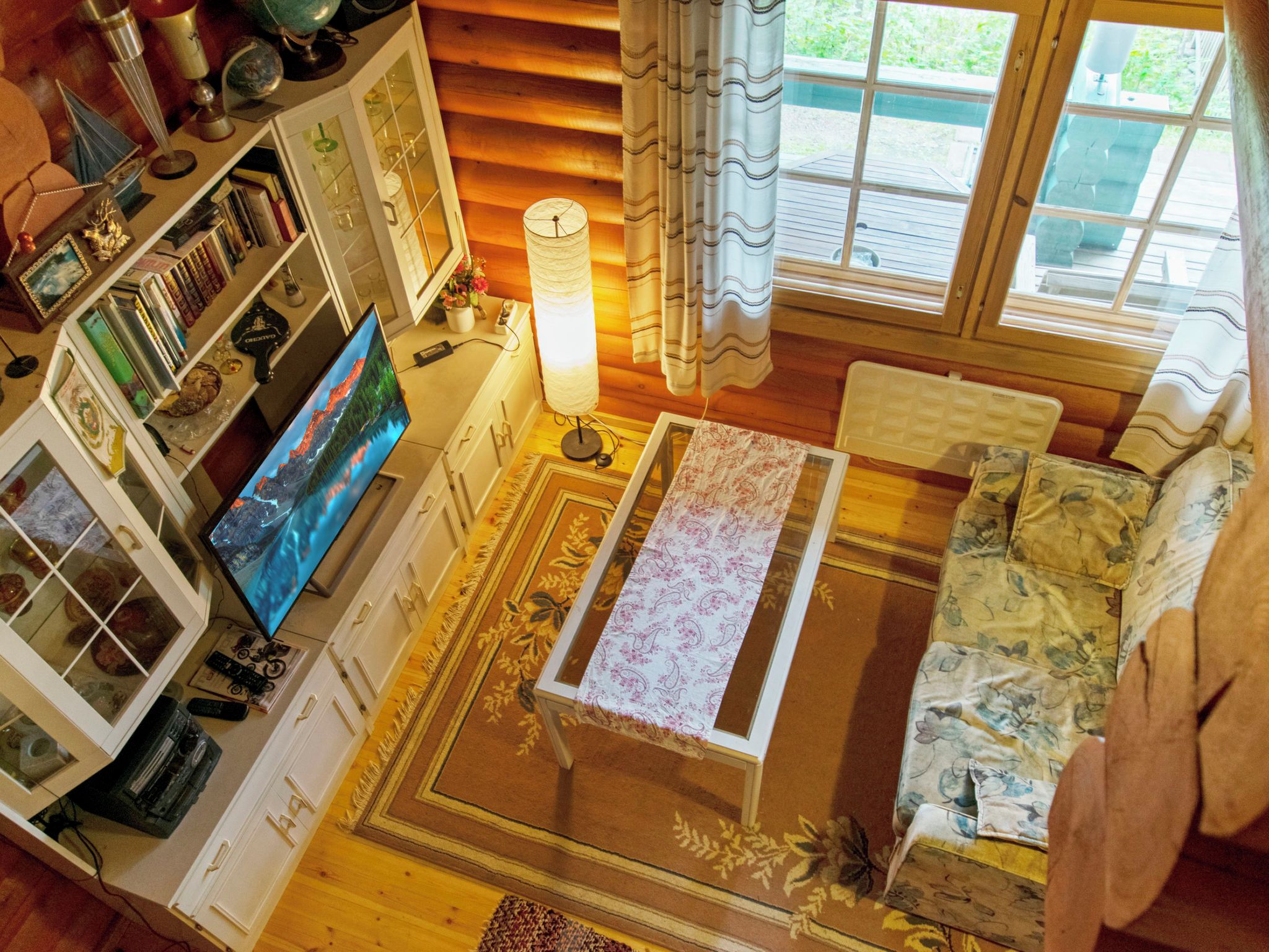 Ein gemütliches Wohnzimmer mit Fernseher, Bücherregalen, einer Lampe und Blick ins Freie.