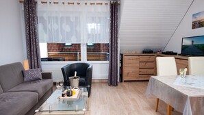 Ein modernes Wohnzimmer mit einer Couch, einem Glastisch, einem Esstisch mit weißer Tischdecke und einem schwarzen Ledersessel.