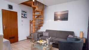 Ein modernes Wohnzimmer mit einer Holztreppe, einem grauen Sofa, einem Glastisch und einem schwarzen Sessel.