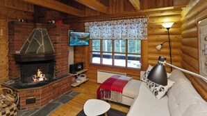 Een knus houten blokhut interieur met een open haard, een televisie, een bank en een raam met uitzicht op bomen.