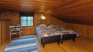 Een gezellig slaapkamer onder het dak, met houten wanden en een schuine hemel. Er staat een bed met quilt, een klein tafeltje met boeken en een raam met uitzicht op bomen.