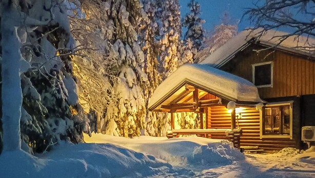 Een houten hutje met een met sneeuw bedekte kap, omgeven door sneeuwzware bomen.