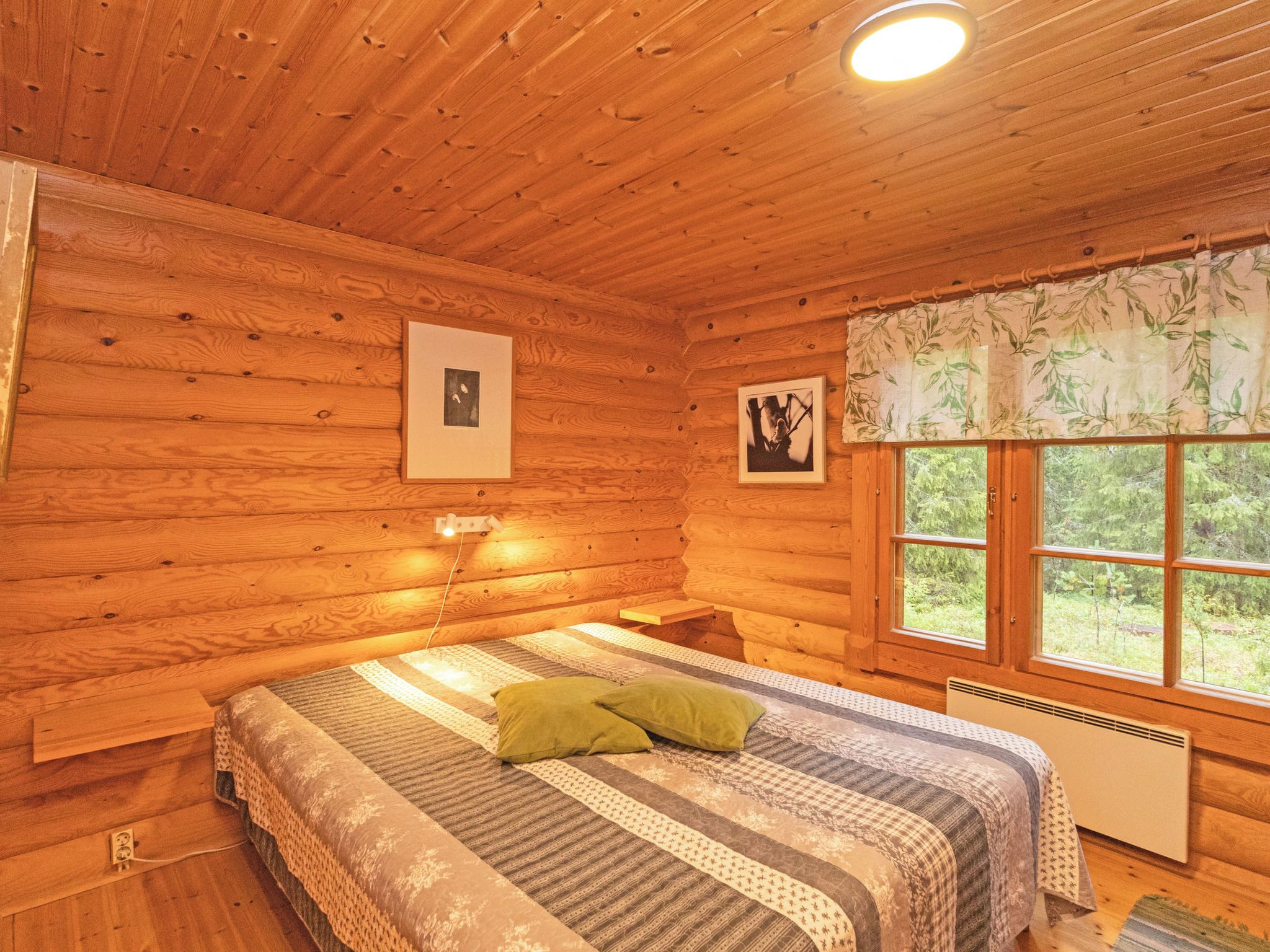 A wooden cabin bedroom with a bed, pillows, framed pictures, a window with a view, and a ceiling light.