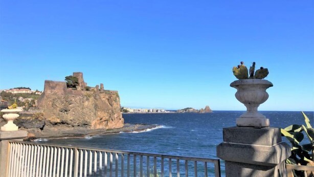 A coastal view with a historic stone structure, a clear blue sky, and a decorative urn on a balcony railing. - Gondar Studio by Interhome (Aci Trezza)