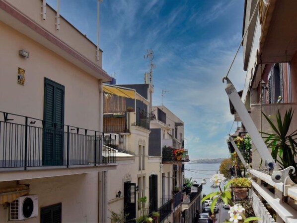 A narrow urban street with buildings on both sides, balconies with plants, and a clear sky.