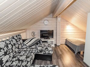 A cozy attic bedroom with a wooden sloped ceiling, a sofa with patterned cushions, a flat-screen TV, and a bed with a blanket.