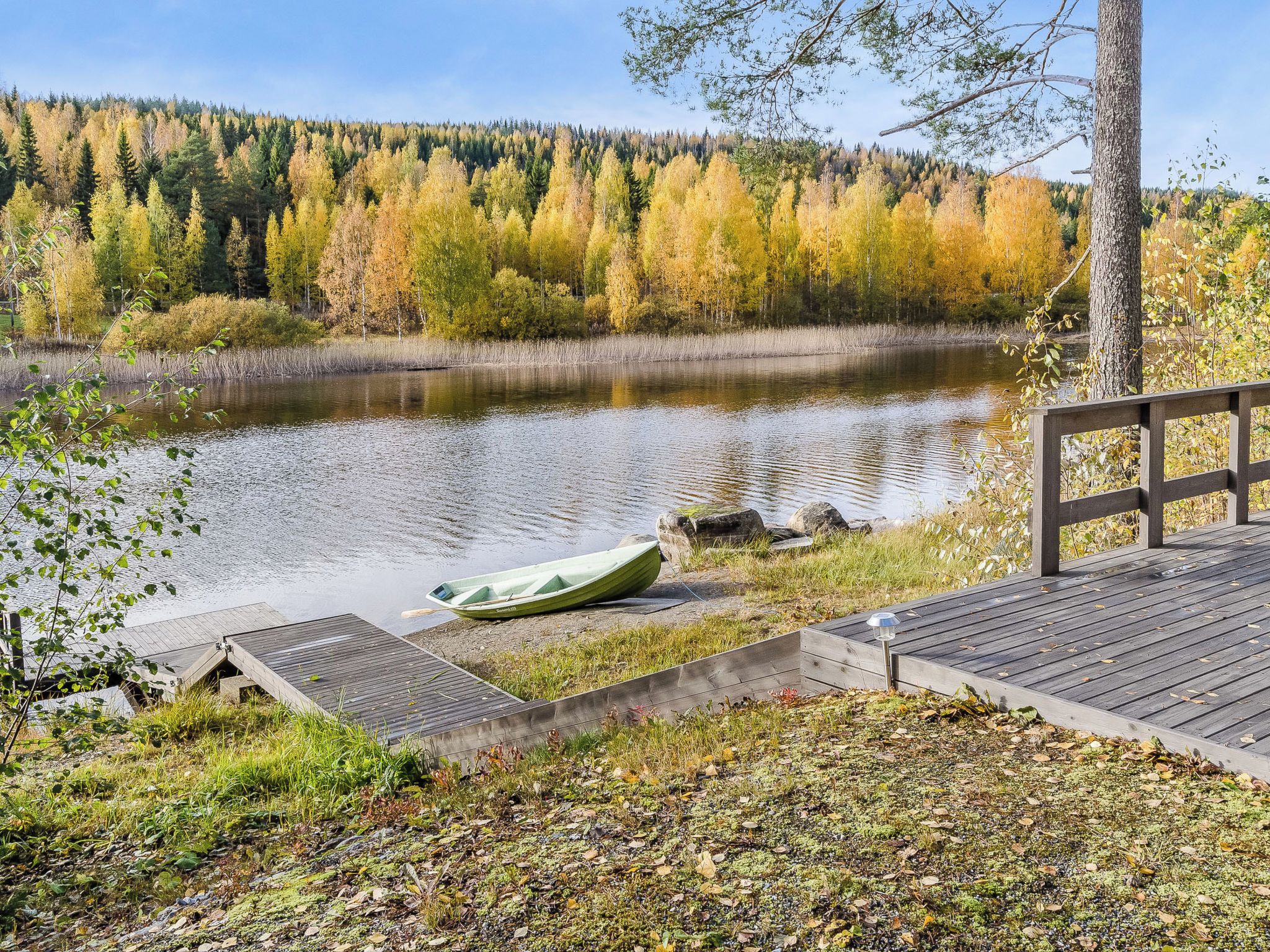 A wooden dock extending over a calm lake with a rowboat, surrounded by autumn-colored trees and a clear sky.