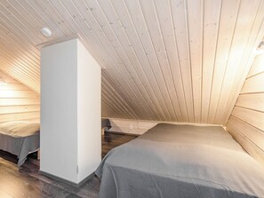 Una habitación pequeña y acogedora con paredes de madera y techo inclinado. Hay una cama con una manta gris y un armario blanco.