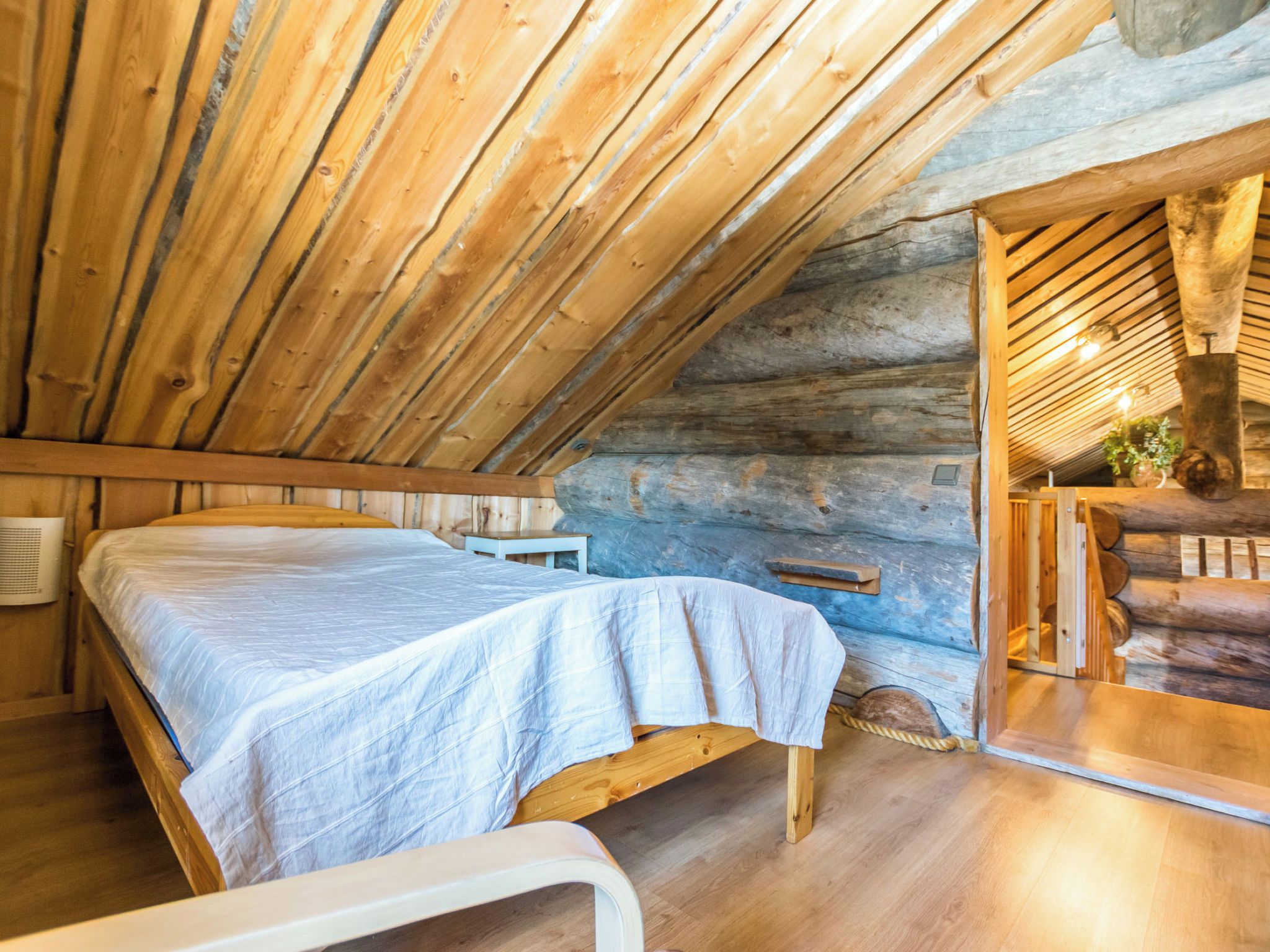 A wooden interior with a sloped ceiling, a bed with a white bedspread, a small table, and a bench.