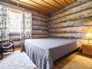 Ein Schlafzimmer in einer Blockhütte mit einem Bett, einem Sessel, einem Nachttisch und einem Fenster mit Vorhängen.
