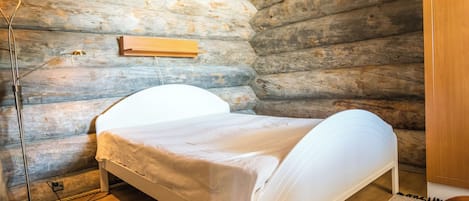 A single bed in a log cabin room with a wooden ceiling and walls, a bedside lamp, and a small wooden shelf.