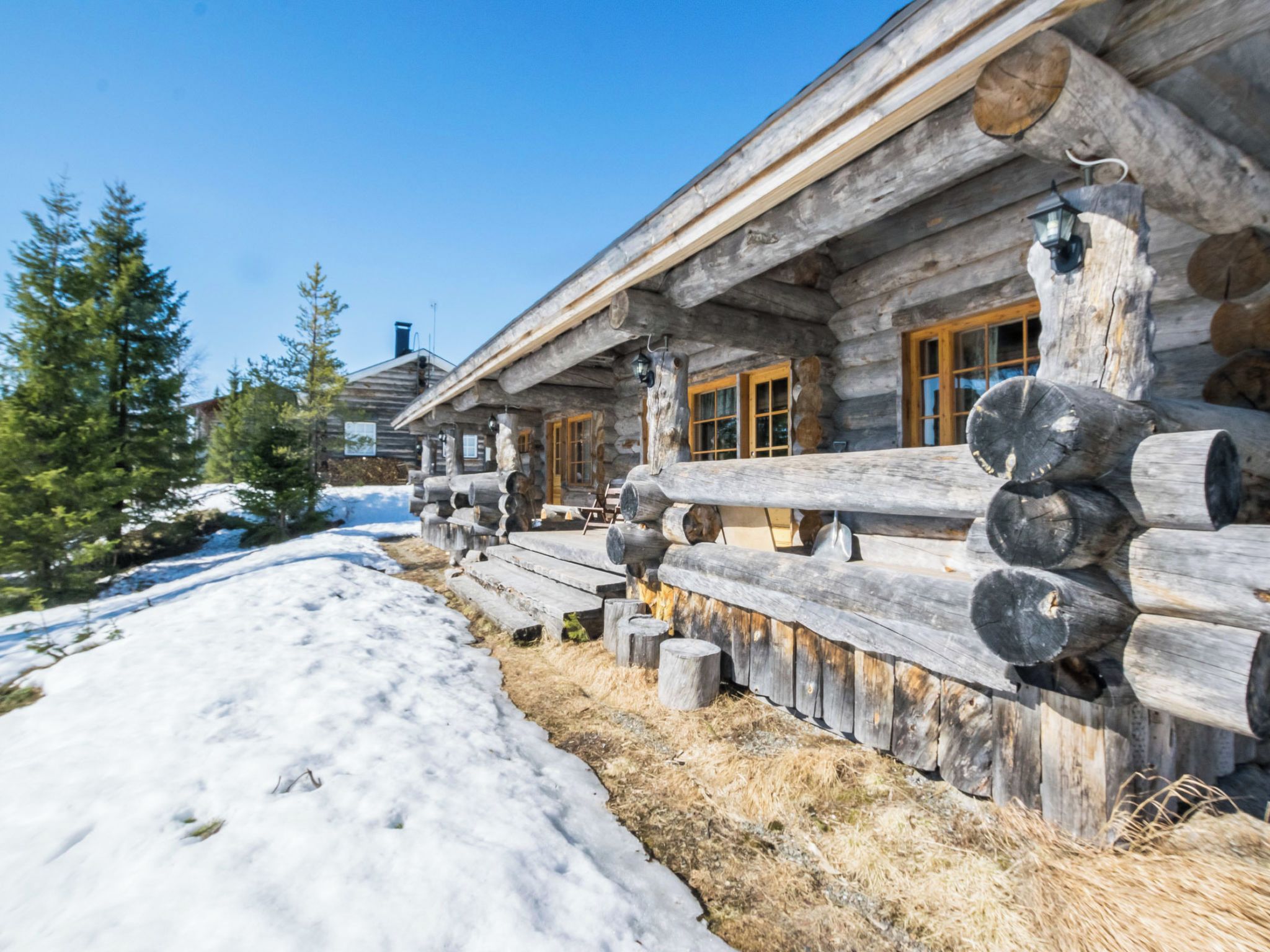 A log cabin with a snow-covered ground, wooden logs stacked outside, and a clear sky.