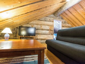 Interior acogedor de una cabaña de troncos con un televisor sobre un soporte de madera, un sofá y una lámpara sobre una mesa.
