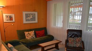 Ein gemütliches Wohnzimmer mit einem grünen Sofa, einem Holztisch und einem Ledersessel. An der Wand hängen zwei gerahmte Bilder.