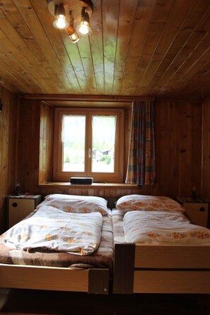 A wooden cabin bedroom with a double bed, two bedside tables, a window with curtains, and a ceiling light fixture. - Talblick by Interhome (Saas-Grund)