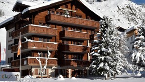 Un edificio in legno a più piani con balconi, circondato da alberi innevati e da montagne sullo sfondo.