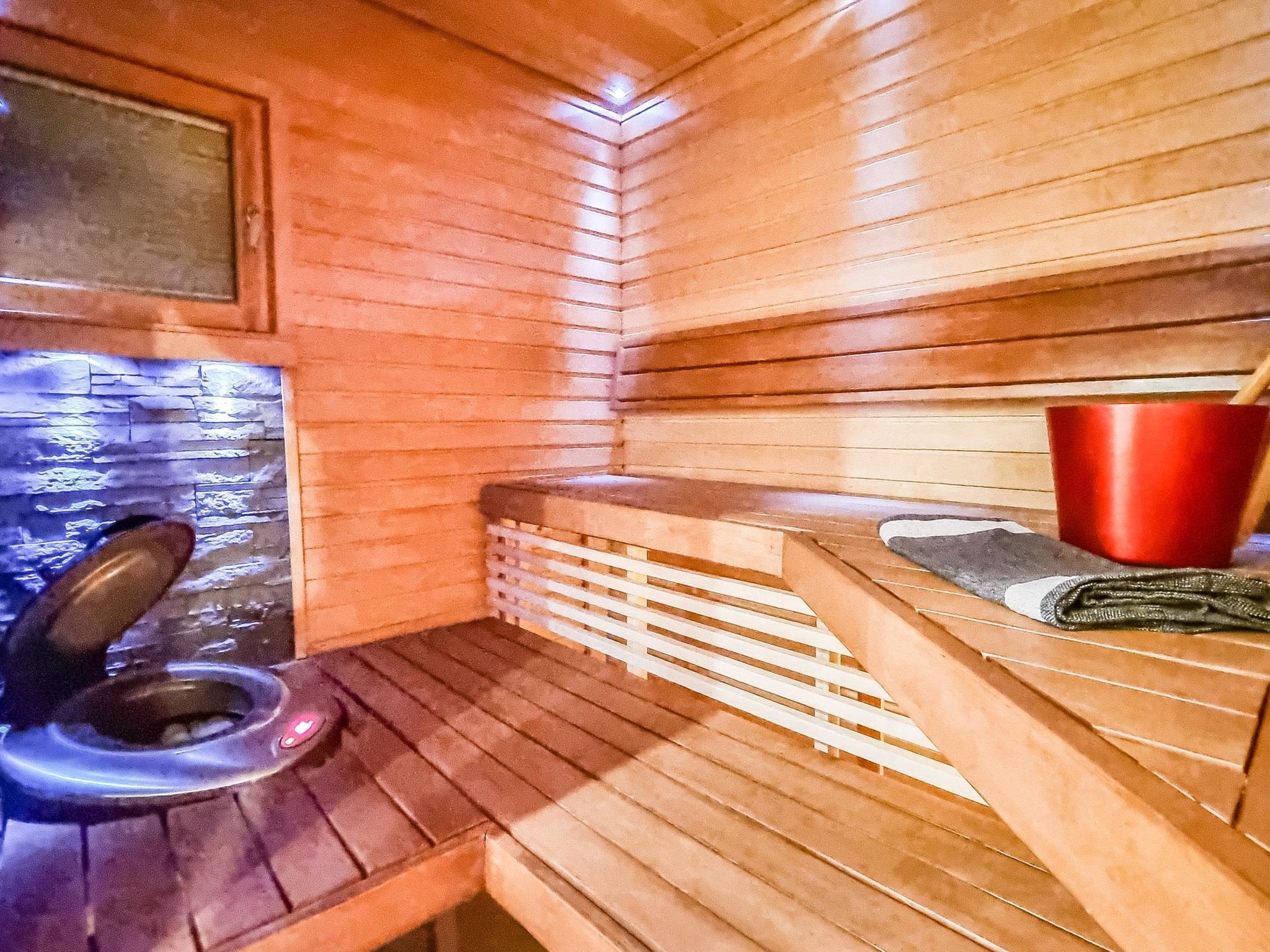 Un sauna avec des bancs en bois, un seau rouge et une fenêtre donnant sur l’extérieur.