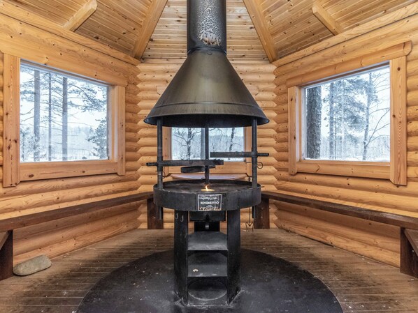 Interior de una cabaña de troncos con un horno de leña en el centro, dos ventanas con vistas nevadas y paredes de madera.
