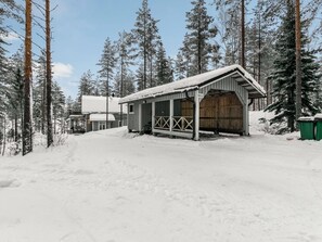 A snow-covered building with a wooden door and a metal roof, surrounded by tall pine trees.