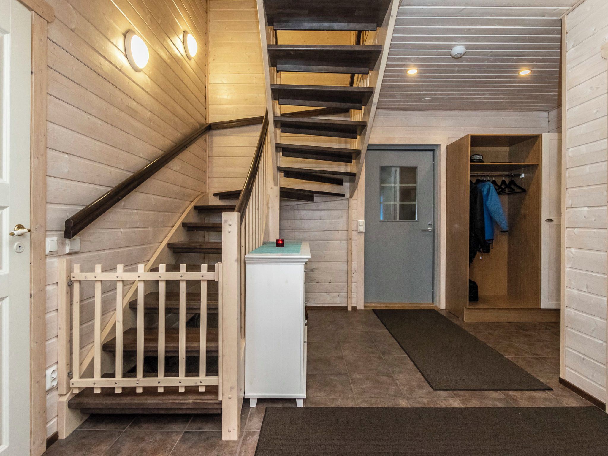 Una escalera de madera que lleva al segundo piso, una puerta con un ventanal y un perchero con ropa colgada.
