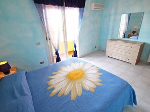 A bedroom with a bed covered in a blue blanket with a large daisy design, a white dresser, a mirror, and a window with curtains.