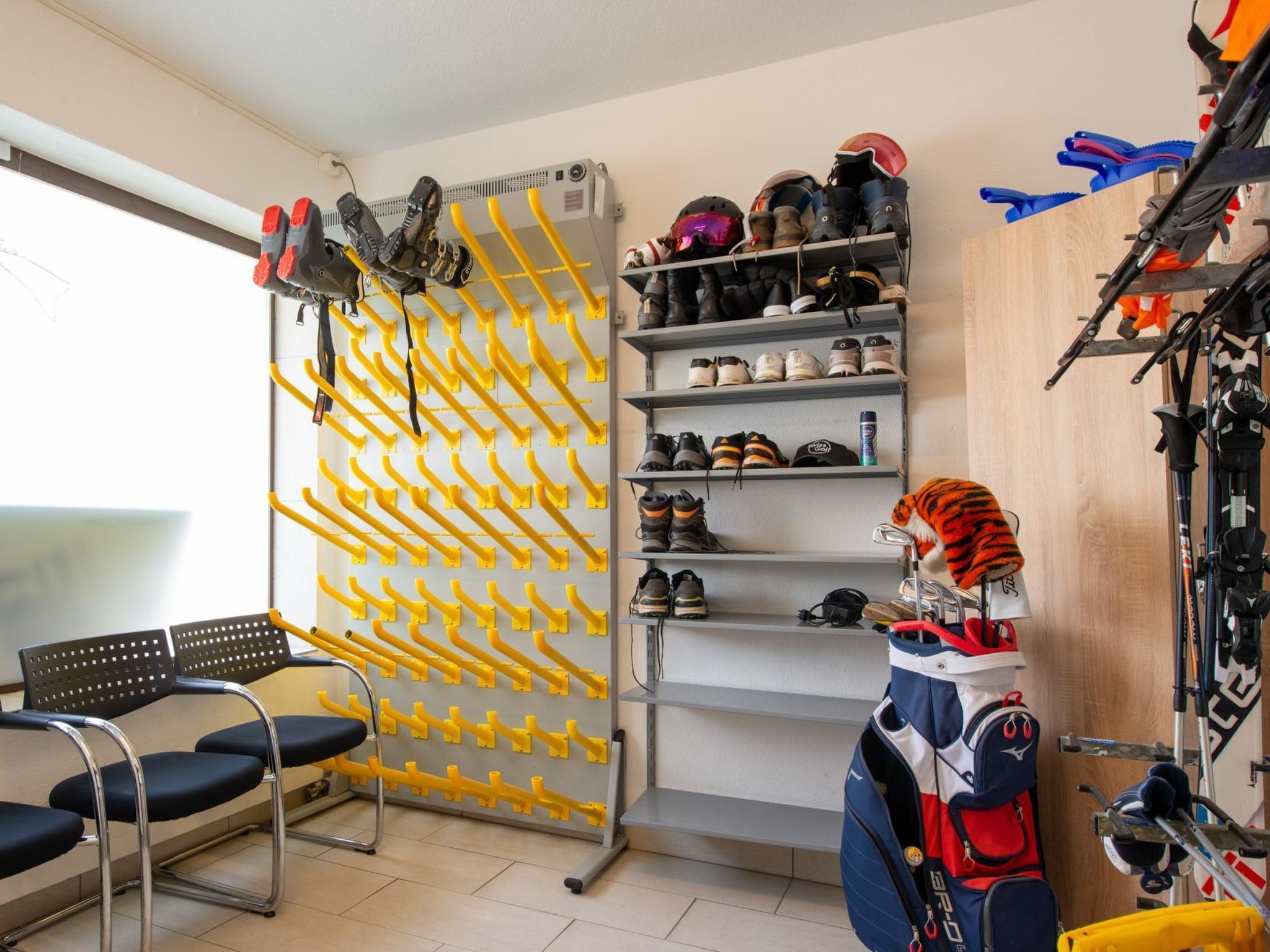 A room with a shoe rack, shelves with helmets and boots, a chair, and a bag.