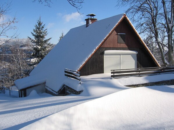 A snow-covered wooden cabin with a sloped roof, a chimney, and a garage door.
