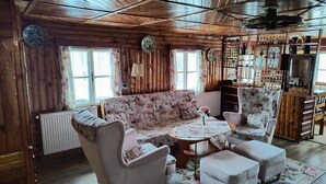 Ein gemütliches Blockhauss-Interieur mit einem floralen Sofa, passenden Sesseln, einem kleinen runden Tisch und einer Kommode mit Dekorationsobjekten.