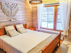 A wooden cabin bedroom with a bed, pillows, a nightstand, a macrame wall hanging, and a window with curtains.
