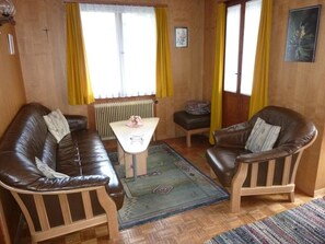 Een gezellige kamer met een bruine leren bank, een klein tafeltje en een stoel. Twee ramen met gele gordijnen, een radiator en een ingelijst kunstwerk op de muur.