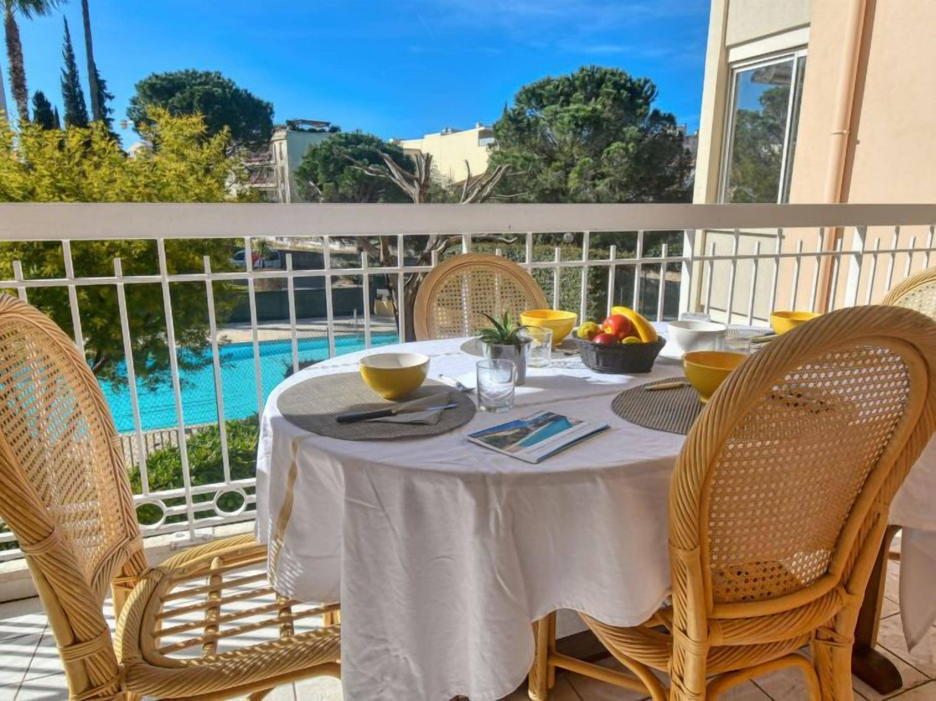 A round table with a white tablecloth, wicker chairs, and a view of a swimming pool.