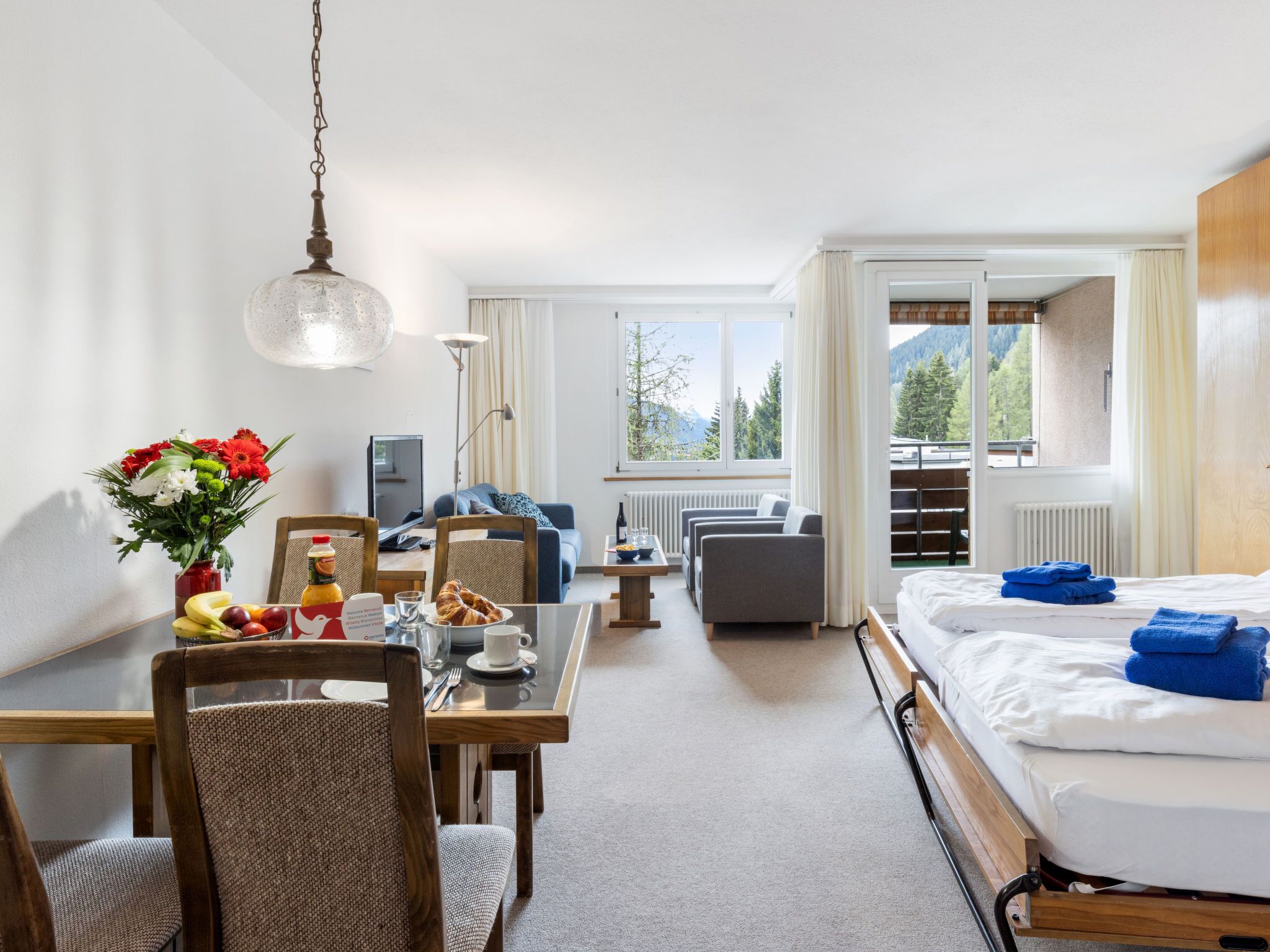 A hotel room with a bed, a dining table with chairs, a sofa, and a view of mountains through the windows.