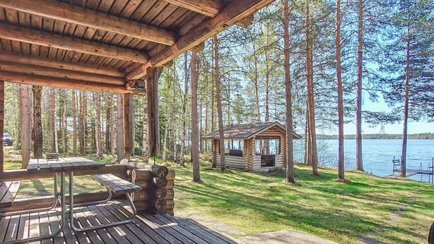 Um deck de madeira com mesas de piquenique e uma cabana pequena junto a um lago, cercado por árvores.