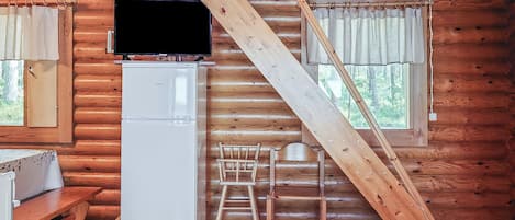 L’intérieur d’une cabane en rondins, avec un téléviseur posé sur un meuble, un réfrigérateur, une chaise en bois et une fenêtre avec des rideaux.