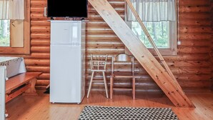 Interior de uma cabana de madeira com um frigorífico branco, uma mesa de madeira, uma cadeira e uma escada de madeira.