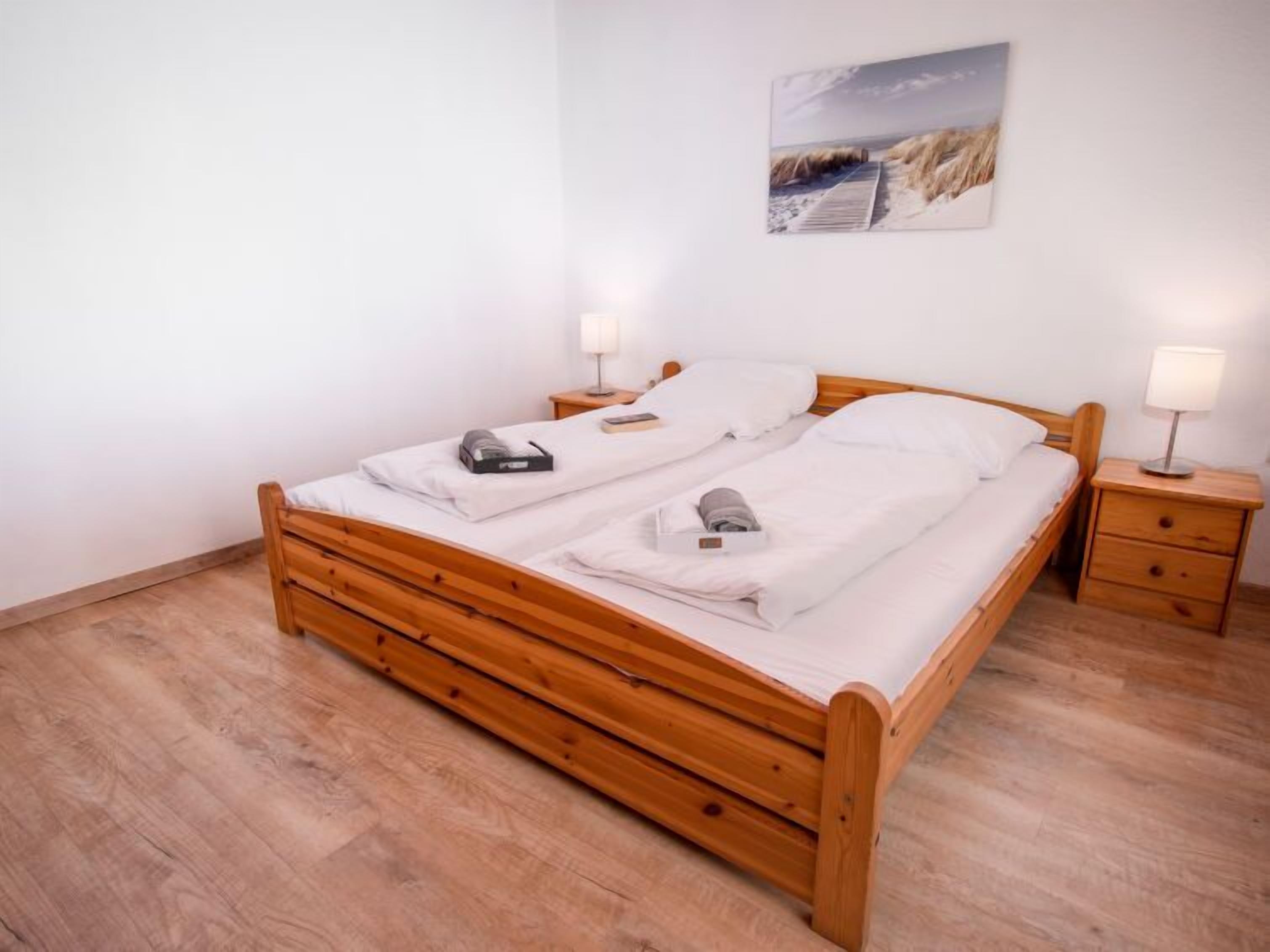 Uma cama de casal de madeira com roupa de cama branca, duas mesas de cabeceira com luminárias e um quadro com tema de praia na parede.