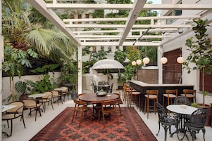 Un espacio exterior cubierto para comer con varias mesas y sillas, rodeado de abundante follaje y una estructura de pérgola blanca.