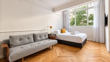 Una habitación de hotel moderna con un sofá gris, una cama con ropa de cama blanca, suelo de madera y una ventana con cortinas.